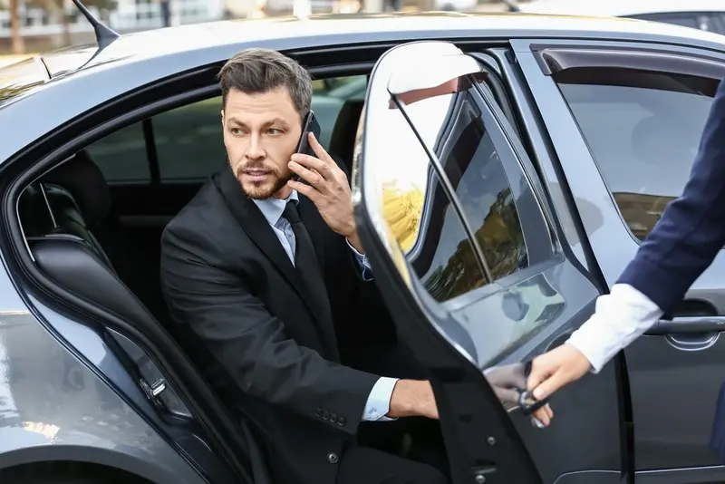 A businessman in a formal suit speaks on his smartphone as he exits a luxurious black car, while a professional driver holds the door open to assist the client in leaving. This symbolizes Houston Town Car Service.